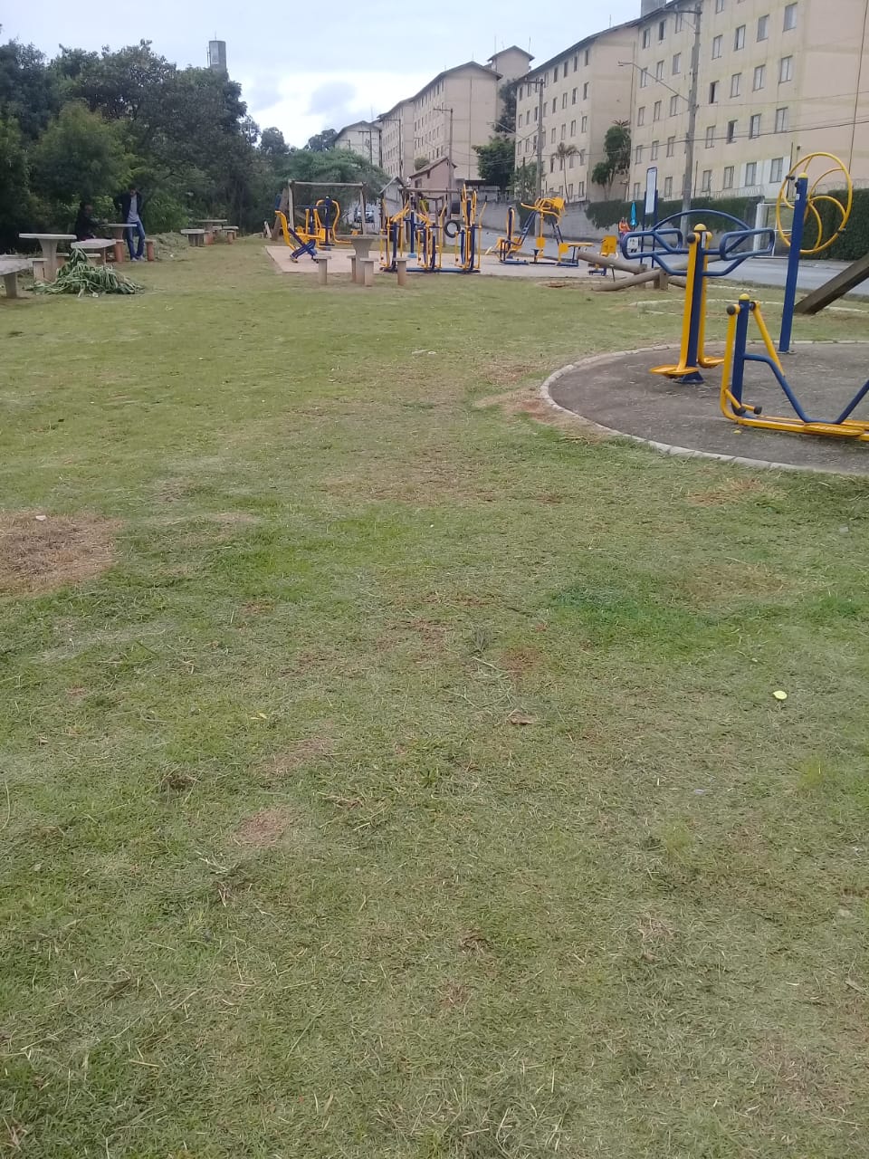 O gramado está limpo e aparado, na rua Emílio Retrosi, no Jardim Marilu. O acesso ao playground, onde a criançada brinca, está seguro, com a grama aparada. No fundo da foto, os prédios de apartamentos populares. Local agradável, com árvores ao redor.  